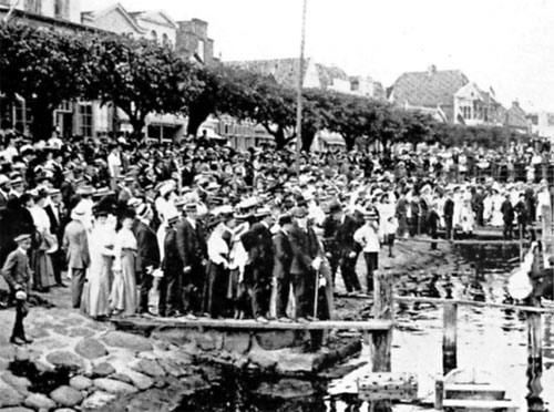 Travemünder Woche 1909 – Die große Hutparade. Mit Melonen und Strohhüten bedeckt werden die Segelyachten von einer große Menschenmenge erwartet.